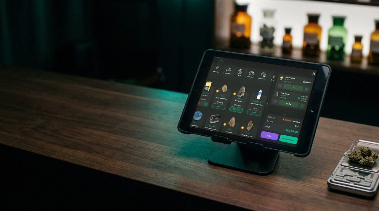 A Budy point-of-sale tablet on a dark wood shop counter, a small bench scale holding flower beside it and shelves of amber apothecary bottles behind.
